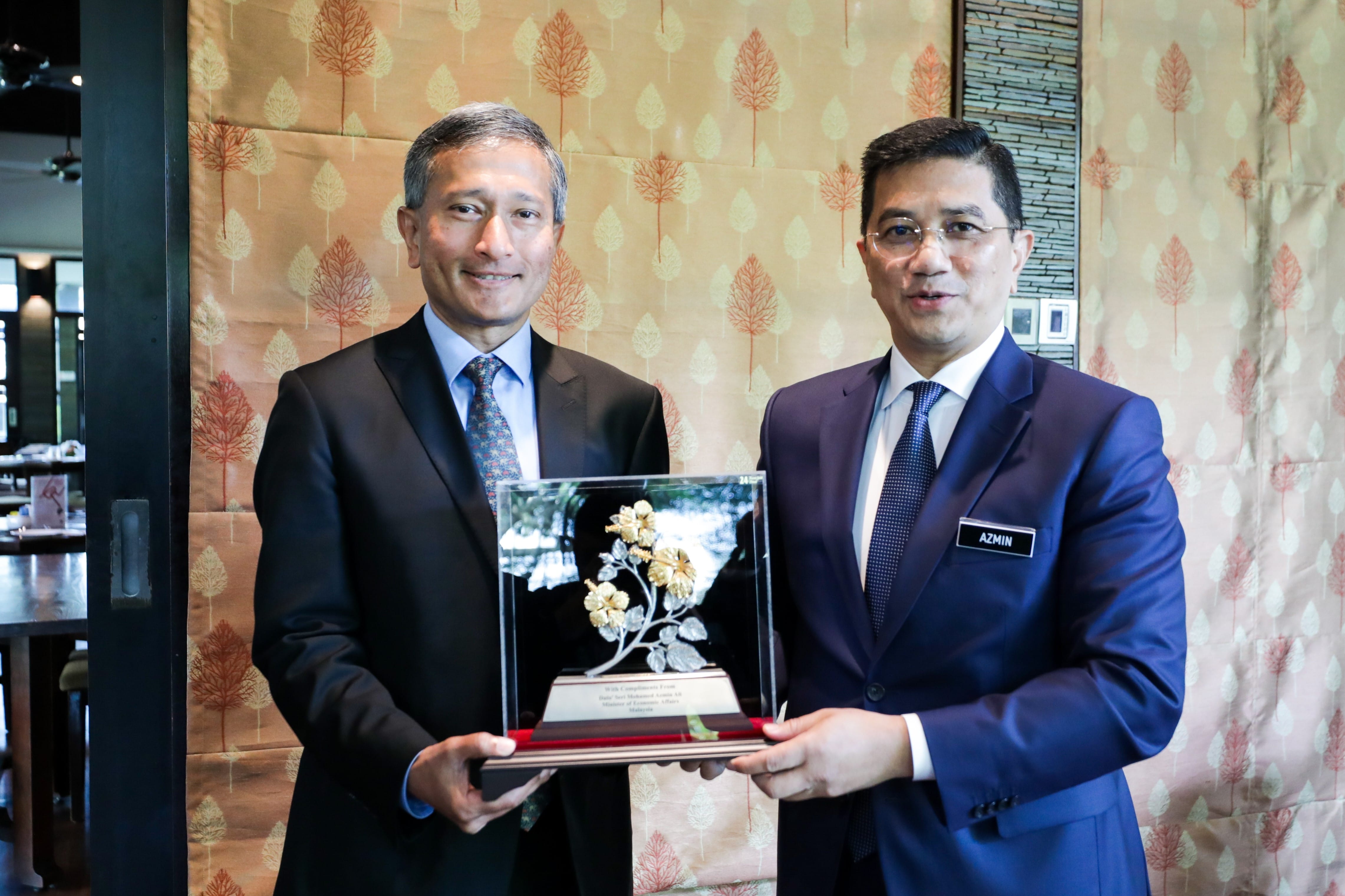 Two men in suits holding a floral award against patterned wallpaper. One man wears "AZMIN" name tag.